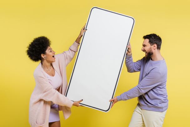 side view of thrilled interracial couple screaming while holding huge phone template isolated on yellow - Foto, imagen