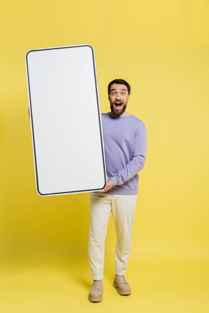 full length of astonished man with open mouth holding huge mock-up of smartphone on grey background - Foto, afbeelding