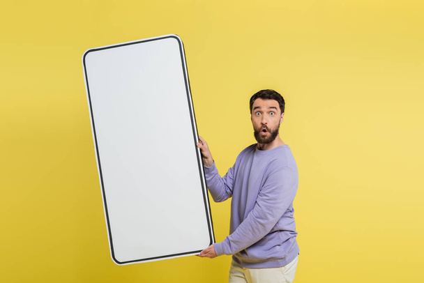 взволнованный бородатый мужчина держит белый шаблон мобильного телефона и глядя на камеру, изолированную на желтый - Фото, изображение