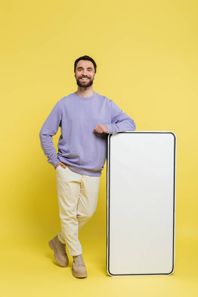 full length of happy bearded man posing with hand in pocket near white mock-up of mobile phone on grey background - Photo, Image