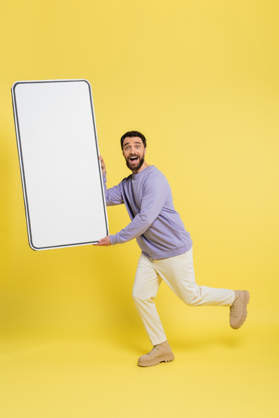 full length of amazed bearded man looking at camera while running with template of cellphone on grey background - Photo, Image