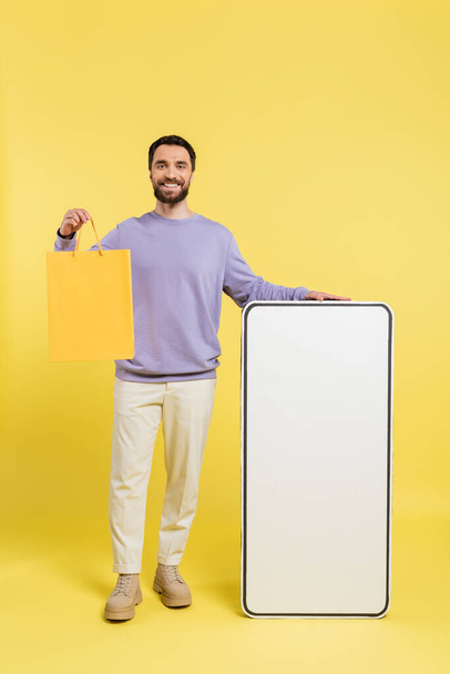 täyspitkä iloinen parrakas mies seisoo ostoskassi ja valtava malli älypuhelimen harmaalla taustalla - Valokuva, kuva
