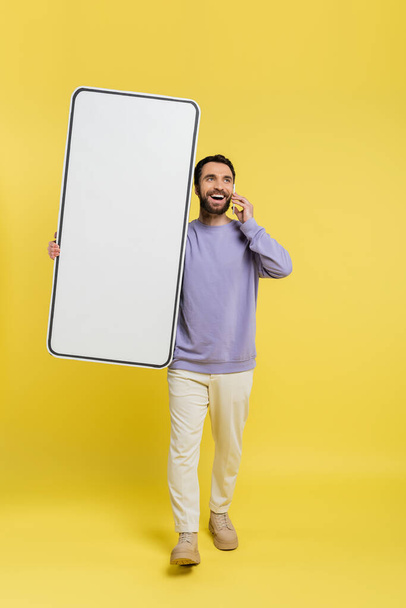 full length of happy man walking with huge phone mock-up and talking on cellphone on grey background - 写真・画像