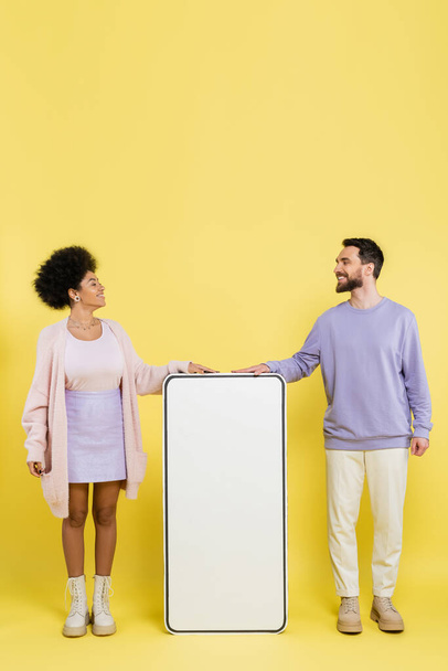 full length on trendy interracial couple smiling at each other near white mock-up of mobile phone on yellow background - Photo, image