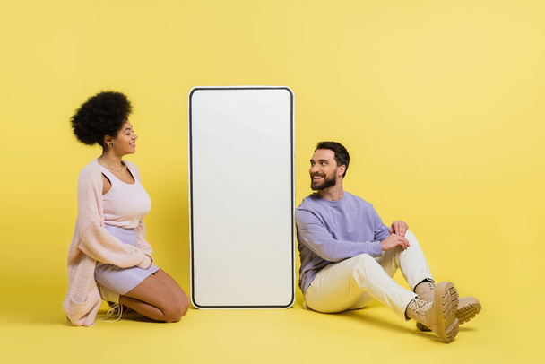 full length on cheerful and trendy interracial couple sitting near white smartphone template on yellow background - Photo, Image