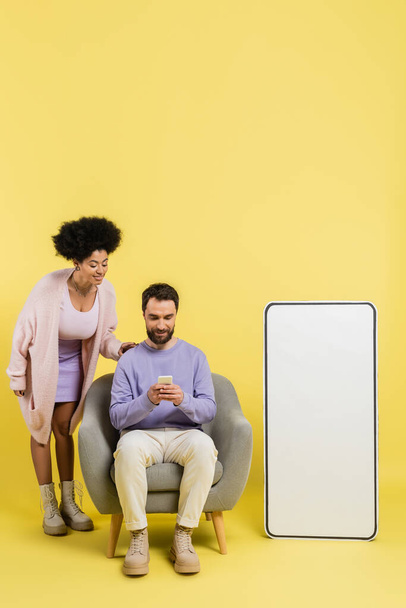 full length of man using smartphone in armchair near smiling african american woman and huge phone template on yellow background - Foto, afbeelding
