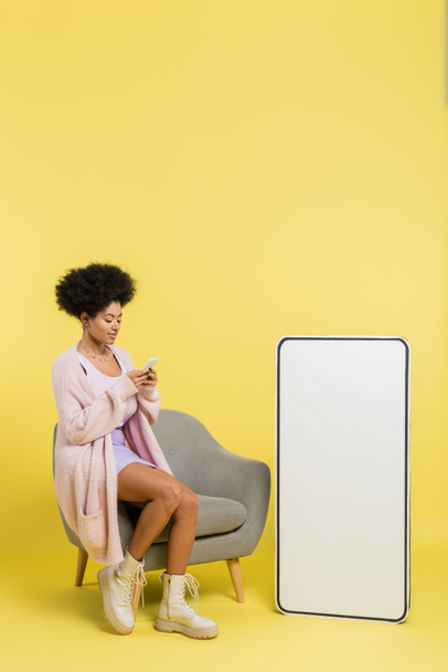 full length of stylish african american woman using mobile phone on armchair near white template of cellphone on yellow background - Photo, Image