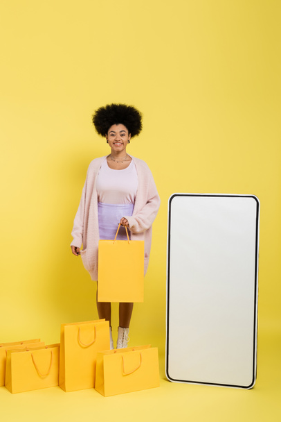 весёлая африканская американка улыбается перед камерой возле торговых мешков и белого шаблона мобильного телефона на жёлтом  - Фото, изображение