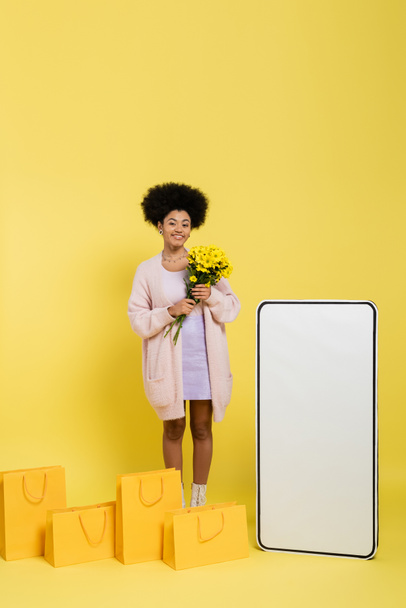 happy african american woman standing with flowers near shopping bags and blank template of smartphone on yellow background - Fotó, kép
