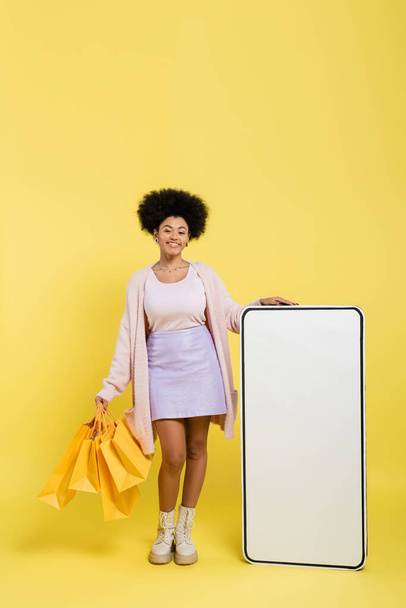 黄色の背景にスマートフォンのカートンモデルの近くにショッピングバッグを持つ喜びと流行のアフリカ系アメリカ人女性の完全な長さ - 写真・画像
