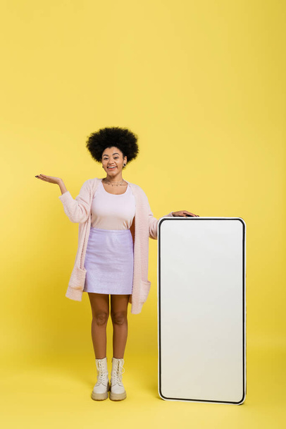 повна довжина веселої афроамериканської жінки, що вказує рукою біля величезного шаблону мобільного телефону на жовтому фоні
 - Фото, зображення