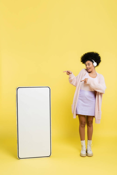 full length of happy african american woman in wireless headphones pointing at empty template of cellphone on yellow background - Valokuva, kuva