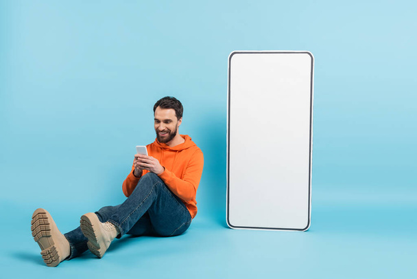 full length of smiling bearded man messaging on smartphone while sitting near huge phone template on blue background - Φωτογραφία, εικόνα