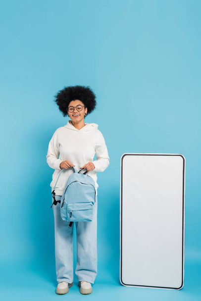 full length of african american student holding backpack and looking at camera near carton phone mock-up on blue background - Photo, image