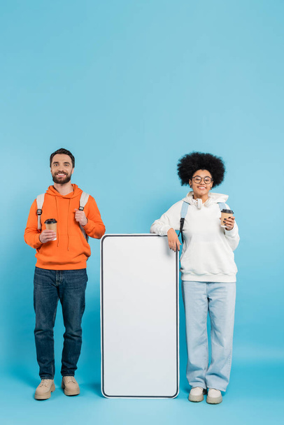 happy interracial students with backpacks and coffee to go near white phone template on blue background - Photo, image