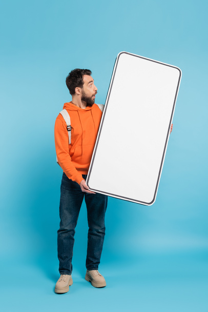 full length of amazed student in orange hoodie holding big phone template on blue background - Foto, imagen