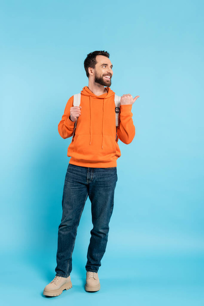 full length of joyful student with backpack looking away and pointing with thumb on blue background - Foto, immagini