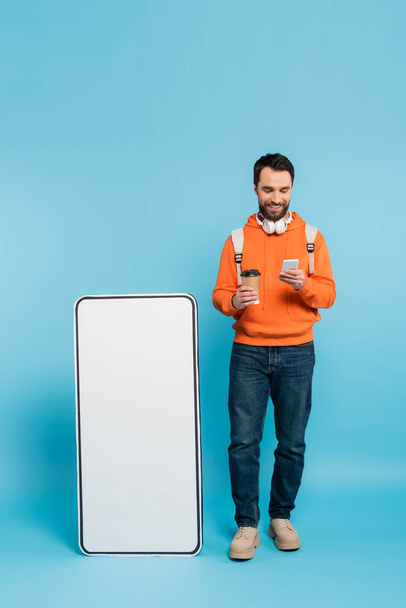 полная длина улыбающийся студент с кофе идти с помощью смартфона возле картонного макета мобильного телефона на синем фоне - Фото, изображение