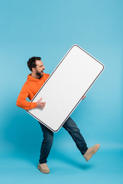 full length of excited man holding huge template of smartphone on blue background - Foto, immagini