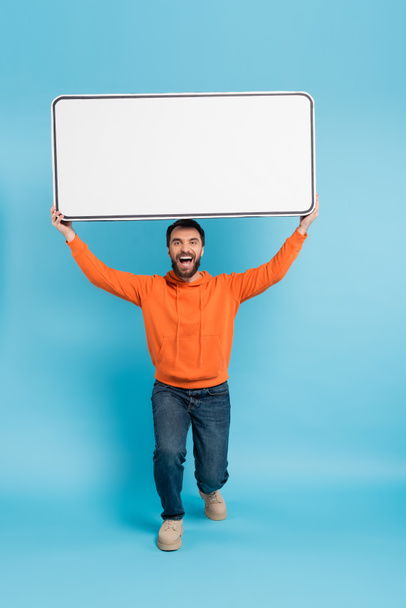 full length of excited man in orange hoodie and jeans holding big phone template above head on blue background - Foto, Imagem