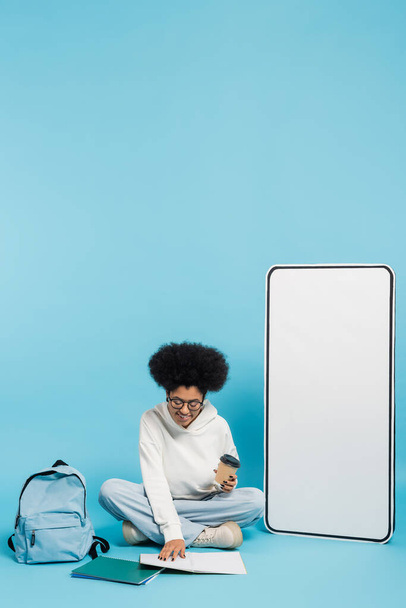 Африканский студент-американец с бумажной чашкой, глядя в блокнот, сидя рядом с рюкзаком и белый телефон макет на синий  - Фото, изображение