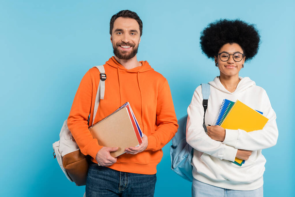 cheerful multiethnic students in hoodies with copybooks and backpacks looking at camera isolated on blue - Photo, Image