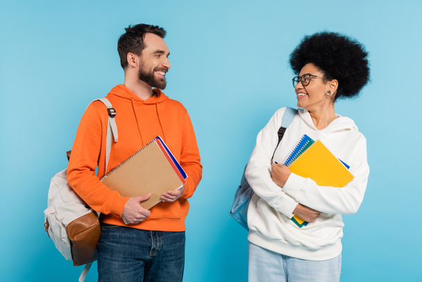 happy and stylish interracial students with backpacks and notebooks looking at each other isolated on blue - Foto, imagen