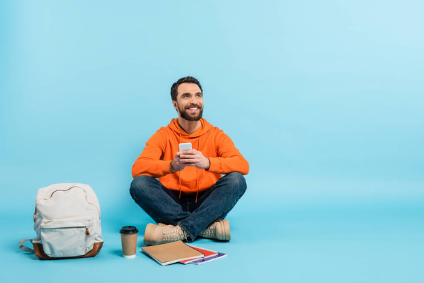 бородатый студент с мобильным телефоном сидит рядом с ноутбуками и рюкзаком, глядя в сторону на синий фон - Фото, изображение