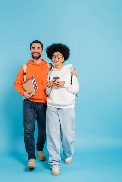 bearded student with notebooks hugging african american girlfriend with paper cup while walking on blue  - Фото, изображение