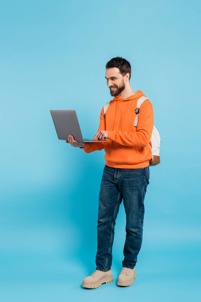 повна довжина щасливого студента в помаранчевому светрі і джинсах, використовуючи ноутбук на синьому
  - Фото, зображення