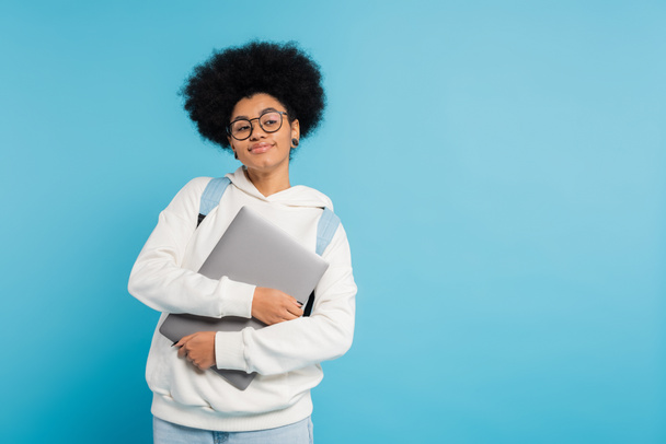 smiling african american student in white hoodie and eyeglasses holding laptop while looking away isolated on blue - Foto, imagen