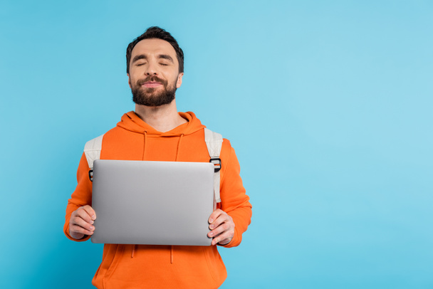 positive student in orange hoodie holding laptop while standing with closed eyes isolated on blue - Foto, Imagem