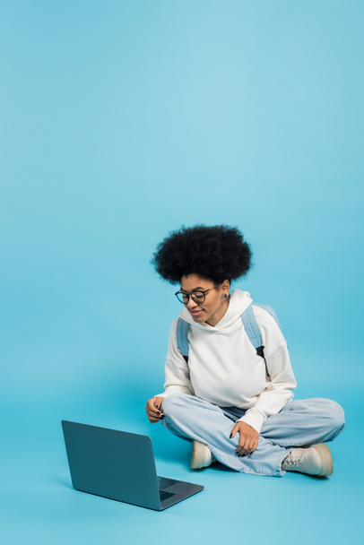 full length of african american student in eyeglasses and white hoodie sitting with crossed legs near laptop on blue background - Valokuva, kuva