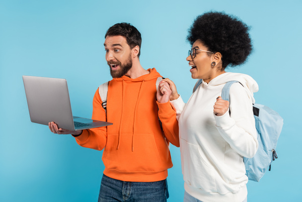 excited interracial students in hoodies looking at laptop isolated on blue - Фото, зображення