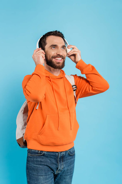 happy bearded student in orange hoodie adjusting wireless headphones isolated on blue - Foto, imagen