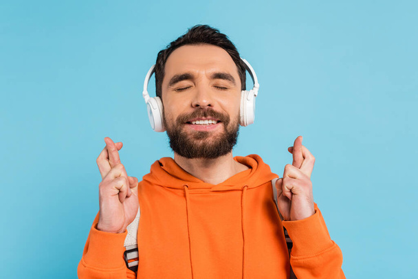 positive student in wireless headphones holding crossed fingers while standing with closed eyes isolated on blue - Foto, Bild