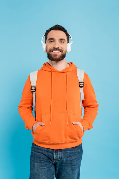 pleased student in wireless headphones and orange hoodie posing with hands in pockets isolated on blue - Fotoğraf, Görsel