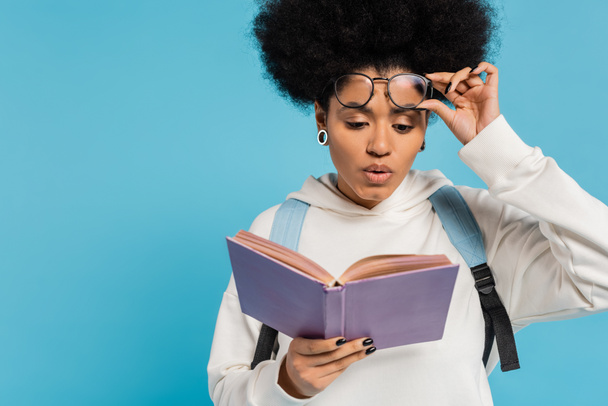 impressed african american student holding eyeglasses while reading book isolated on blue - Foto, Imagem
