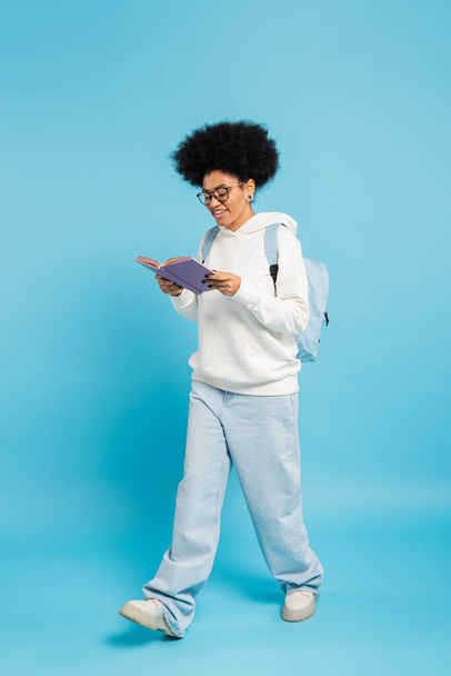 full length of smiling african american student in eyeglasses and jeans reading book while walking on blue  - Valokuva, kuva
