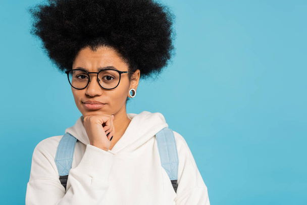 upset and thoughtful african american student in eyeglasses holding hand near chin and looking at camera isolated on blue - Photo, Image