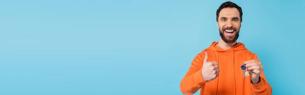 excited brunette man in orange hoodie showing like gesture while holding key isolated on blue, banner - Foto, imagen