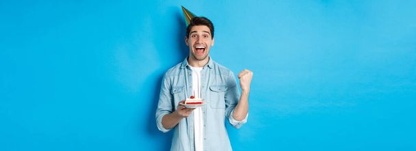 Veselý muž vypadá šťastně, slaví narozeniny ve slavnostní čepici, drží b-den dort a dělat pěst čerpadlo, stojící nad modrým pozadím. - Fotografie, Obrázek