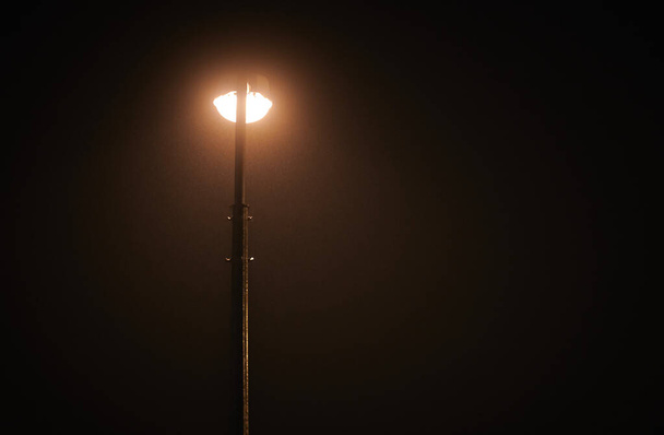 One night lamppost shines with faint mysterious yellow light through evening fog. Streetlight shine at quiet city night, magic atmospheric light in mystical darkness, right copy space - Φωτογραφία, εικόνα