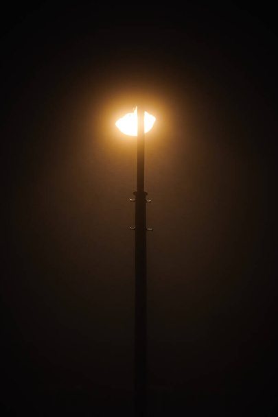 One night lamppost shines with faint mysterious yellow light through evening fog. Streetlight shine at quiet city night, magic atmospheric light in mystical darkness - Zdjęcie, obraz