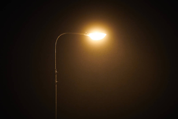 One night lamppost shines with faint mysterious yellow light through evening fog. Streetlight shine at quiet city night, magic atmospheric light in mystical darkness, copy space - Foto, afbeelding