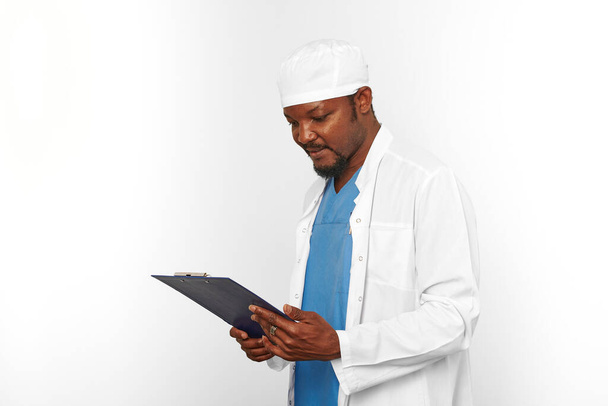 Black surgeon doctor bearded man in white coat and cap looks to medical chart on clipboard, isolated on white background copy space. Pensive adult black african american practicing surgeon portrait - Foto, Bild