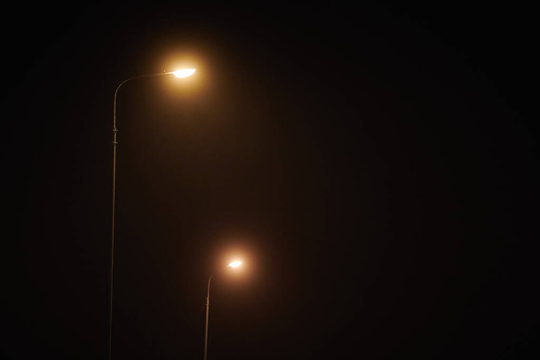 Two night lampposts shines with faint mysterious yellow light through evening fog. Streetlights shine at quiet city night, magic atmospheric light in mystical darkness - Φωτογραφία, εικόνα