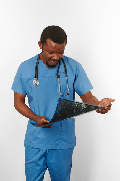 Black surgeon doctor bearded man in blue coat with stethoscope holds X ray image, isolated on white background. Serious black african american practicing radiologist portrait, confident look to xray - Photo, Image