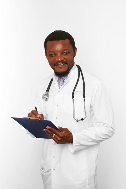 Smiling black bearded doctor man in white robe with stethoscope filling medical records on clipboard, isolated on white background. Happy adult black african american physician therapist portrait - Photo, Image