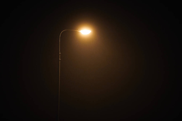 One night lamppost shines with faint mysterious yellow light through evening fog. Streetlight shine at quiet city night, magic atmospheric light in mystical darkness, copy space - Foto, Imagen